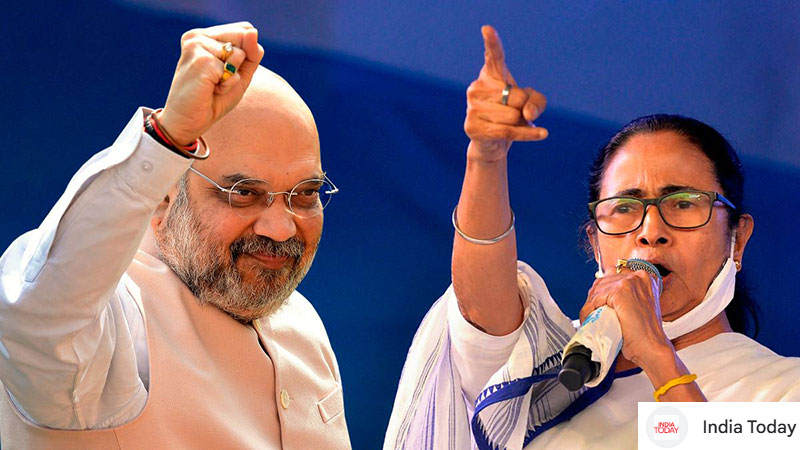 Two speakers at a rally on stage; a bearded man with glasses raises a fist while a woman speaks into a microphone and points upward against a blue backdrop.